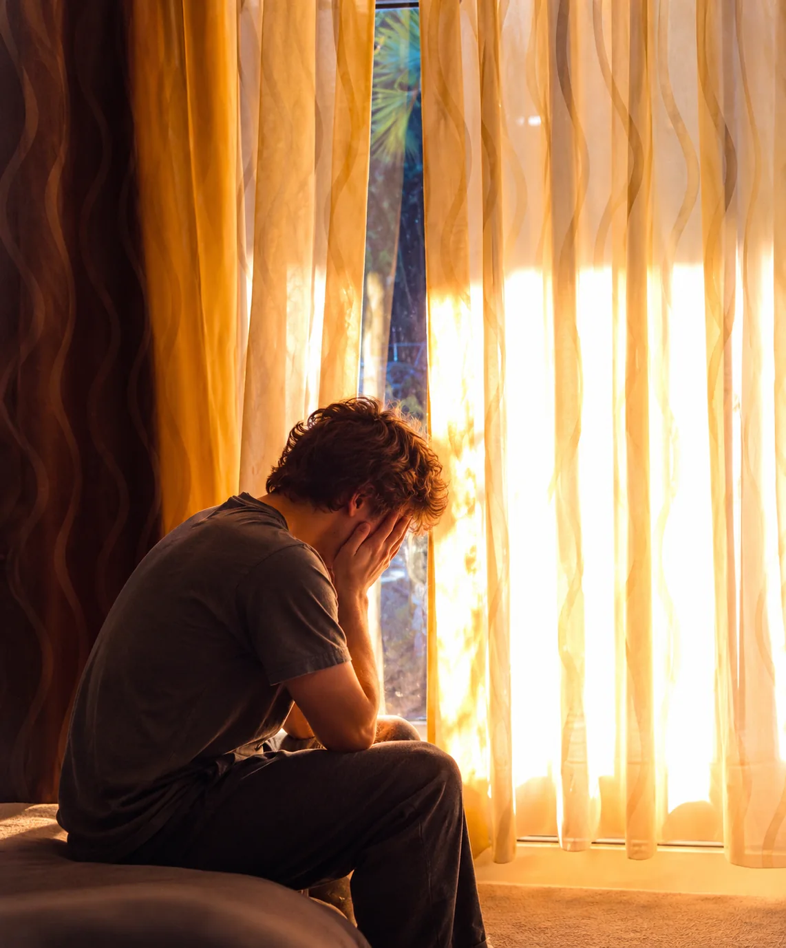 Person sitting at a glowing window with face concealed in hands